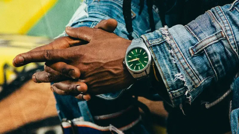 Close-up of clasped hands wearing a green-faced watch with distressed denim jacket against colorful background.