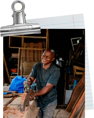 A smiling carpenter planing a plank in a workshop, pinned to a lined index card with a bulldog clip