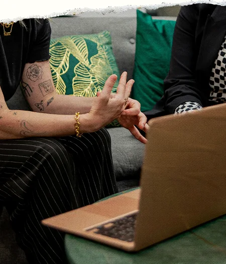 Two people talking on a couch with green cushions and an open laptop, with a torn paper top edge