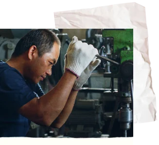 A machinist in work gloves operating a lathe, with a crumpled sheet of paper tucked behind the photo