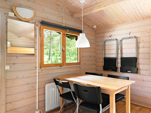 Enkel matplats med ett träbord, fyra svarta stolar och två hopfällbara stolar på en vägg med ljus träpanel; fönstret visar grönska utanför.