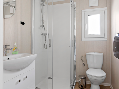 Ett litet badrum med en duschvägg i glas, ett vitt handfat, en väggspegel, en toalett, ett litet fönster och en tvålpump på handfatet.