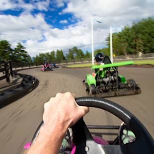 Förstapersonsvy av en person som kör en gokart på en utomhusbana och följer andra gokarts i en kurva under en delvis molnig himmel.