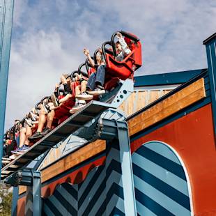 People riding a red roller coaster with their hands up, passing over a blue and red geometric-patterned structure on a sunny day.