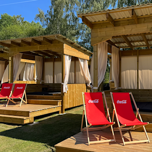 Två röda Coca-Cola-däckstolar står på en träplattform nära cabanas med gardiner, under en klarblå himmel och omgivna av träd.