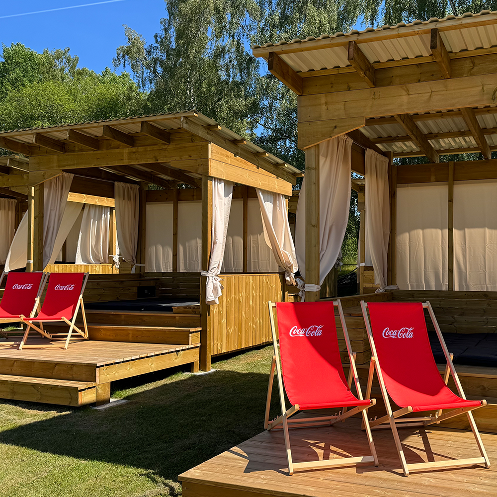 Två röda Coca-Cola-däckstolar står på en träplattform nära cabanas med gardiner, under en klarblå himmel och omgivna av träd.