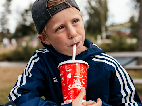 En pojke i mörkblå jacka och bakvänd keps dricker ur en röd mugg med sugrör utomhus.