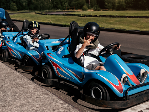 Två barn med hjälmar sitter i blå gokarts som är parkerade på en bana, och det ena barnet håller upp ett fredstecken och ler mot kameran.