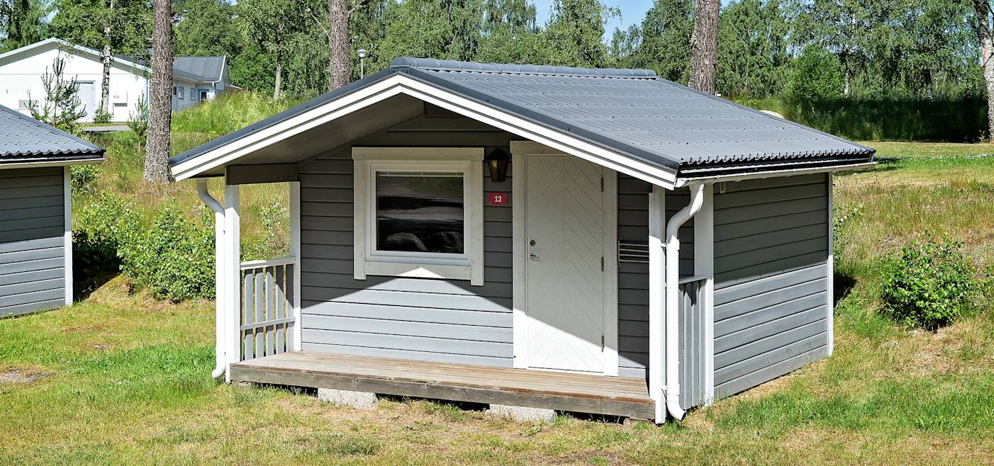 A small gray wooden cabin with a white door and window, a covered front porch, and a metal roof, situated on a grassy area with trees in the background.
