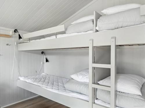 White bunk beds with neatly arranged bedding, attached reading lamps, and a wooden ladder in a small, minimalist room with white paneled walls and ceiling.
