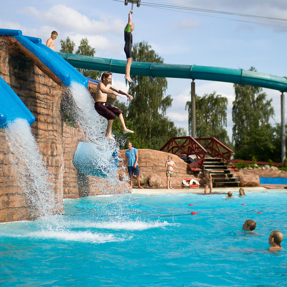 Barn hoppar i en pool från en klippkonstruktion med vattenfall i en vattenpark, medan andra simmar och leker i vattnet nedanför.