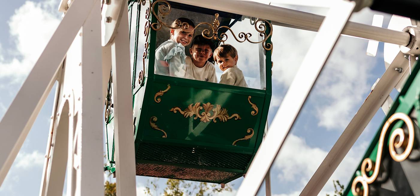Tre barn sitter i en grön pariserhjulsgondol och tittar ut och ler. Himlen och en del av pariserhjulets struktur syns i bakgrunden.