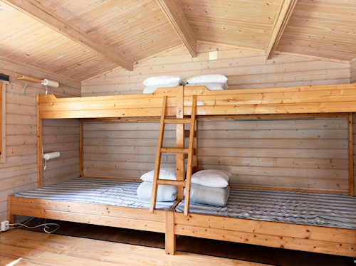 Våningssängar i trä med randiga madrasser och kuddar i ett ljust rum med träpanel. En trästege är placerad i mitten för åtkomst till den översta våningssängen.