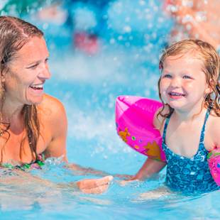 En vuxen kvinna och en ung flicka med flytringar ler när de leker tillsammans i en swimmingpool.