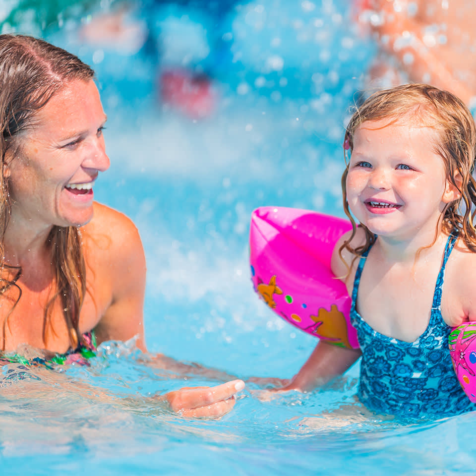 En vuxen kvinna och en ung flicka med flytringar ler när de leker tillsammans i en swimmingpool.
