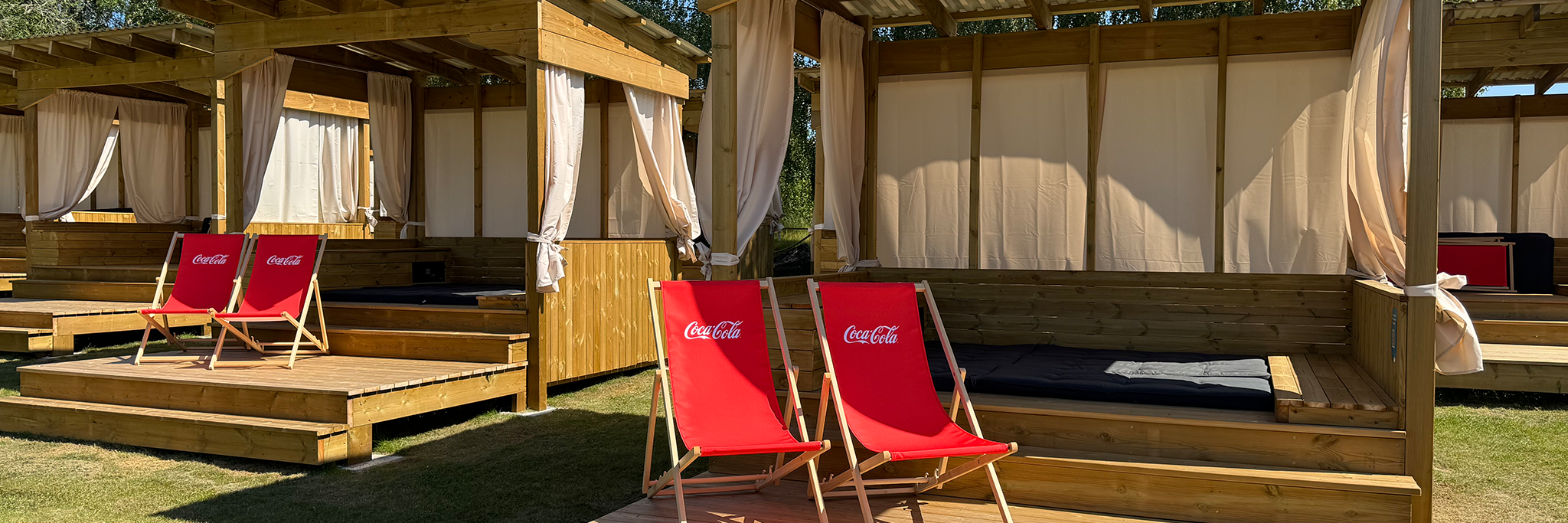 Flera träcabanas med beige gardiner och två röda Coca-Cola solstolar på en gräsyta i solljus.