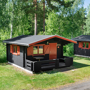 En liten svart trästuga med orangefärgade detaljer och en täckt veranda står på en gräsmatta omgiven av träd och grönska.