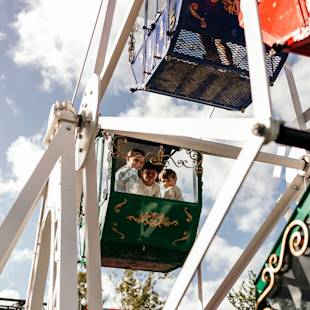 Three children ride in a green Ferris wheel gondola, smiling at the camera, with a blue sky and clouds in the background.