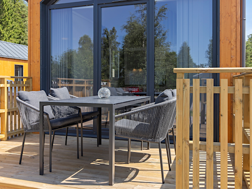 En modern uteplats med ett svart matbord och fyra dämpade stolar på ett trädäck, framför stora glasdörrar och omgiven av ett träräcke.