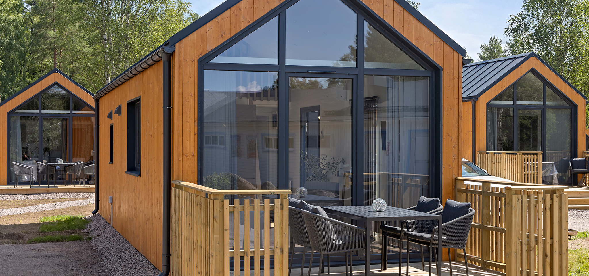 Moderna trästugor med stora glasfönster och små uteplatser, var och en möblerad med bord och stolar, i en grön, trädkantad miljö.