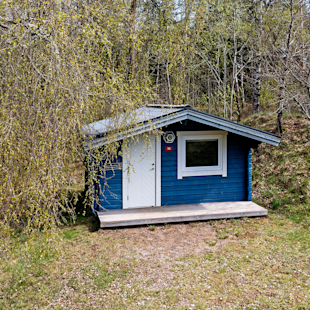 En liten blå trästuga med vit dörr och fönster står i en gräsbevuxen glänta omgiven av träd.