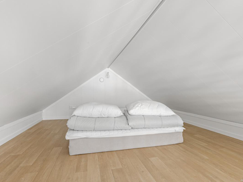 En låg säng med vita sängkläder är placerad under ett snett vitt tak i ett minimalistiskt vindsrum med ljust trägolv.