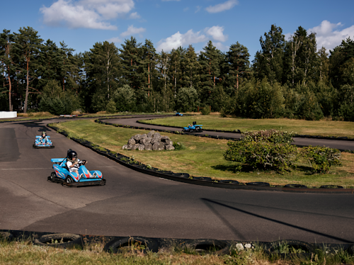 Fyra personer kör blå gokart på en slingrande utomhusbana omgiven av träd och grönska under en blå himmel med spridda moln.