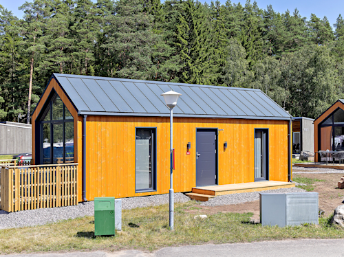 Ett modernt, litet gult hus med mörkt plåttak och stora fönster står på en grusad tomt, omgiven av träd och elskåp.