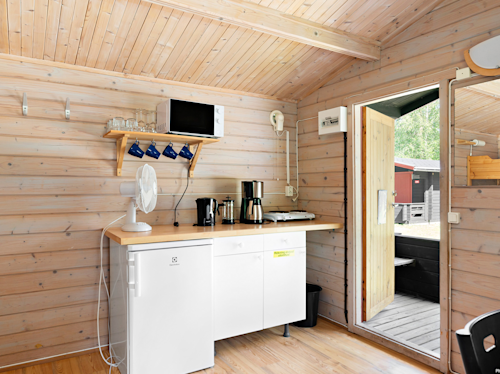 Litet pentry i trä med bänkskiva, vitt kylskåp, mikrovågsugn, kaffebryggare, koppar, glas på en hylla och en öppen dörr som leder ut.