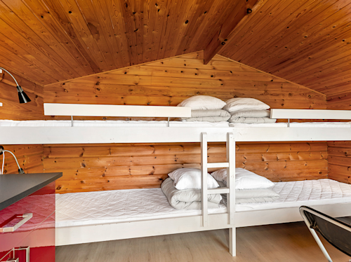 I ett trärum med snedtak finns vita våningssängar, var och en med en madrass, hopvikta sängkläder och kuddar; en svart skrivbordslampa och en del av en röd disk syns till vänster.