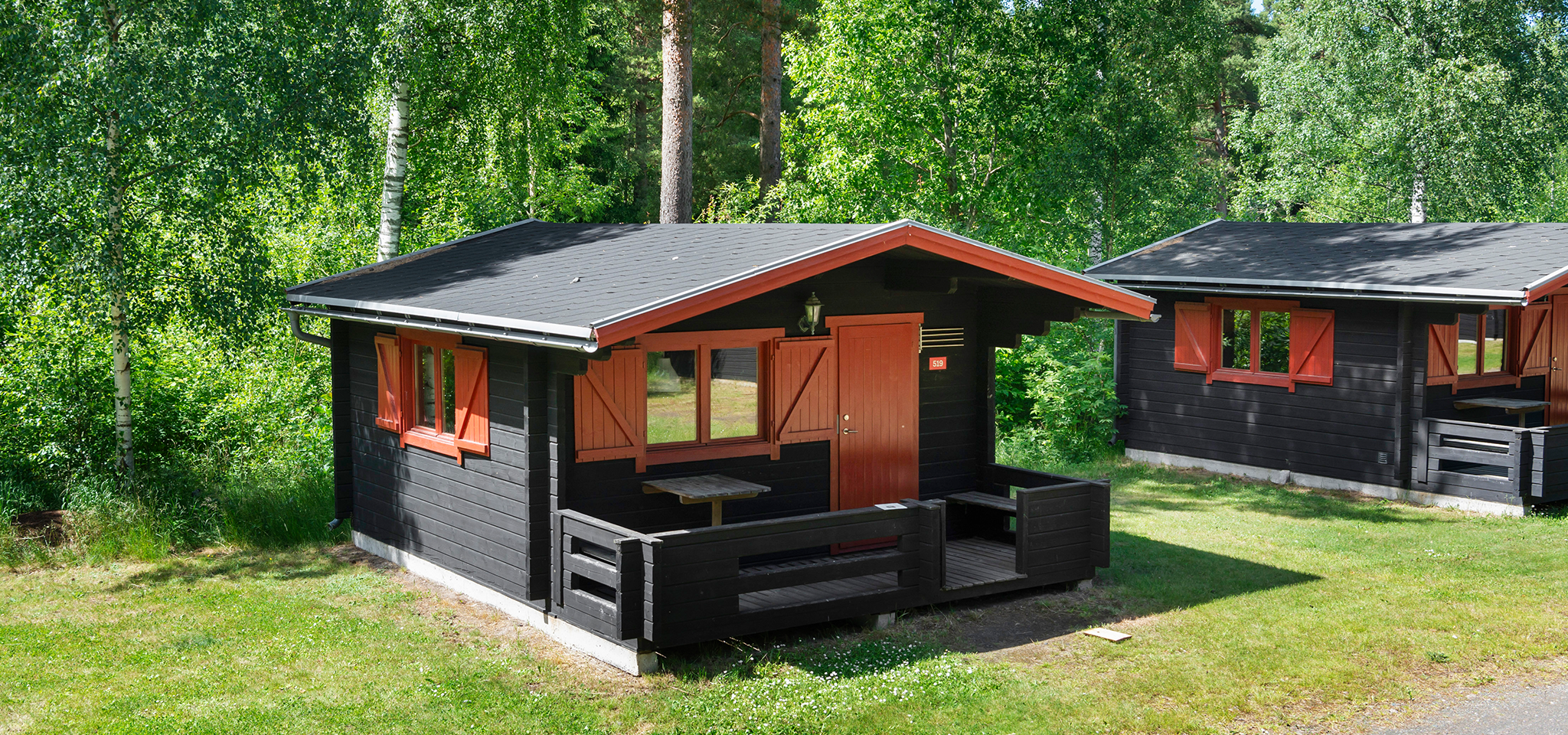 På en gräsmatta omgiven av gröna träd ligger en liten svart trästuga med röda detaljer och en veranda med bord och bänk.