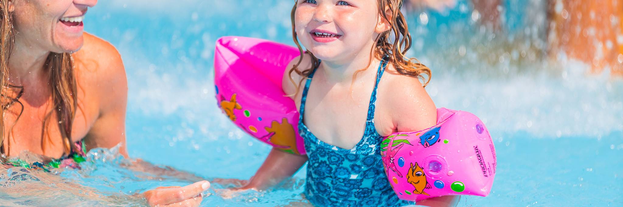 Ett litet barn med rosa flytringar ler när hon leker i en pool med en vuxen bredvid sig.