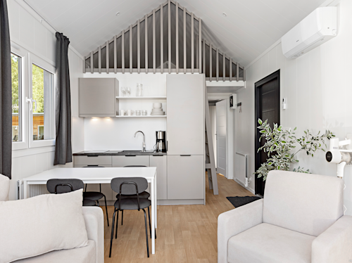 Modernt, minimalistiskt kök och vardagsrum med neutrala toner, ett litet matbord, två stolar, ljusa soffor och en dekorativ växt under ett snedtak.