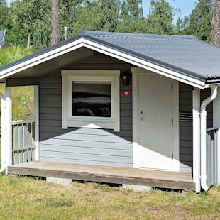 Small gray wooden cabin with a white door and window, porch railing, and a sloped metal roof, surrounded by grass and trees.