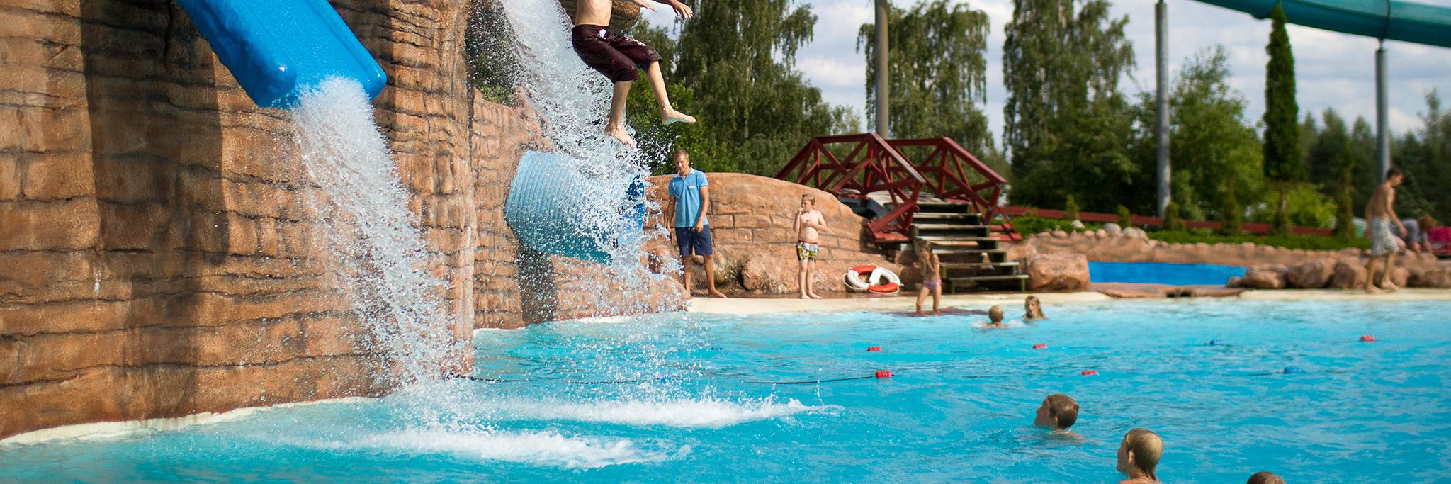 En person glider nerför en blå vattenrutschkana och plaskar ner i en pool medan andra tittar på och leker i vattnet i en utomhusvattenpark.