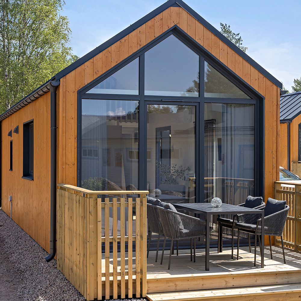 Ett modernt litet hus med träfasad, stora glasfönster och ett litet utedäck med bord och fyra stolar.