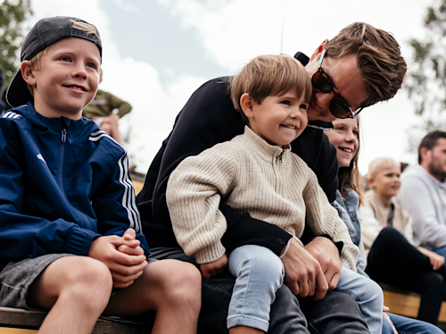 En man sitter nära en liten pojke i sitt knä, omgiven av andra barn och vuxna, som alla verkar titta på något utomhus.