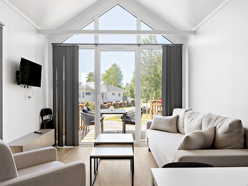 Modernt vardagsrum med vita väggar, beige soffor, en väggmonterad TV och stora fönster med svarta vertikala persienner, med utsikt över en uteplats och en trädgård med träd och ett annat hus utanför.