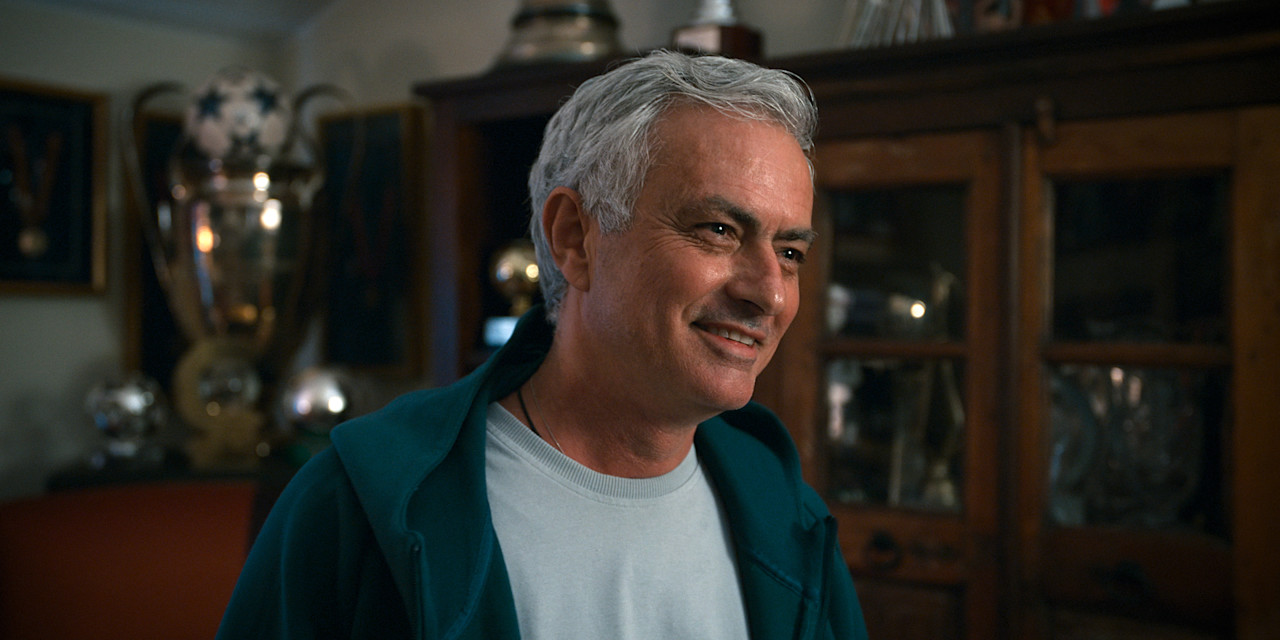 José Mourinho de sorriso aberto, em camisola branca e casaco azul-petróleo, sentado em frente a uma vitrina cheia de troféus
