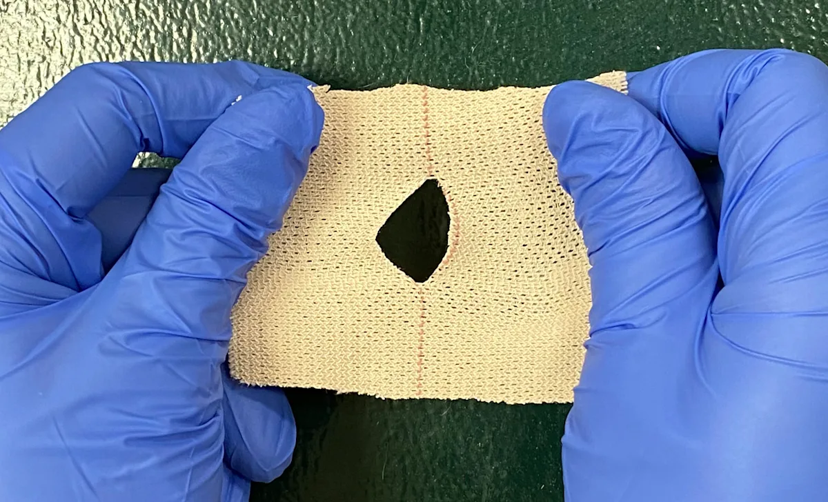 Gloved hands holding elastic tape with hole cut in center and bandaged leg shown with elastic tape placed on end of bandage.