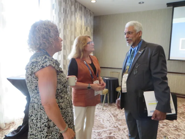Two people attentively watching while third person with laptop case and papers under arm speaks with them.