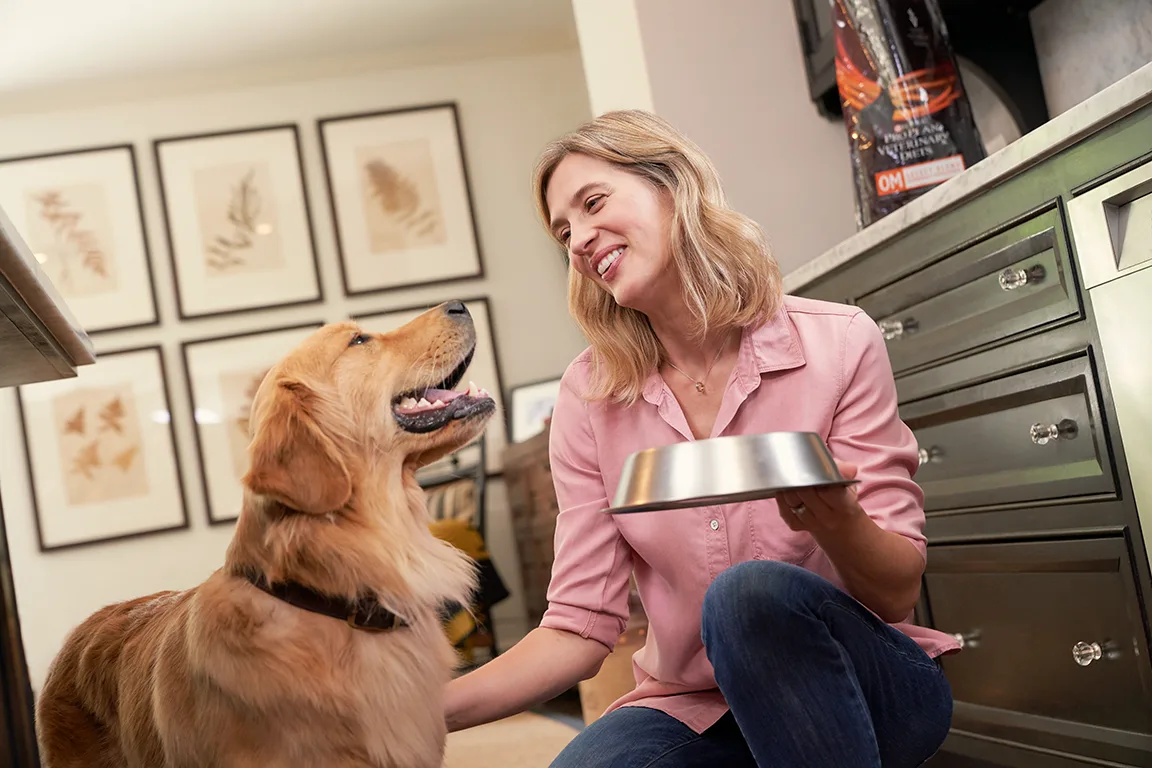 A woman kneels beside a golden retriever in a home setting, smiling as she holds a metal food bowl. The dog looks up at her with a happy expression, while framed botanical prints adorn the wall in the background.