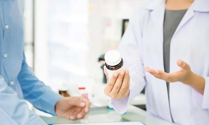 A veterinarian in a white lab coat holds a bottle of medication while discussing it with a client, who is wearing a blue shirt. The setting appears to be a veterinary clinic, with various medical supplies visible in the background.