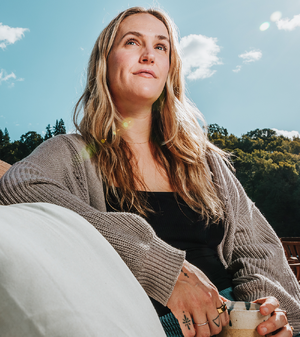 Rachael Brooke Hoppock is sitting on a wooden deck holding a warm beverage, against a peaceful outdoor backdrop.