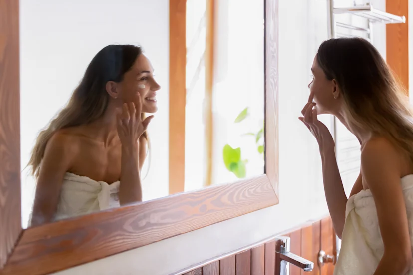 Woman wrapped in a towel looking in the mirror and applying hydroquinone underneath her eyes.