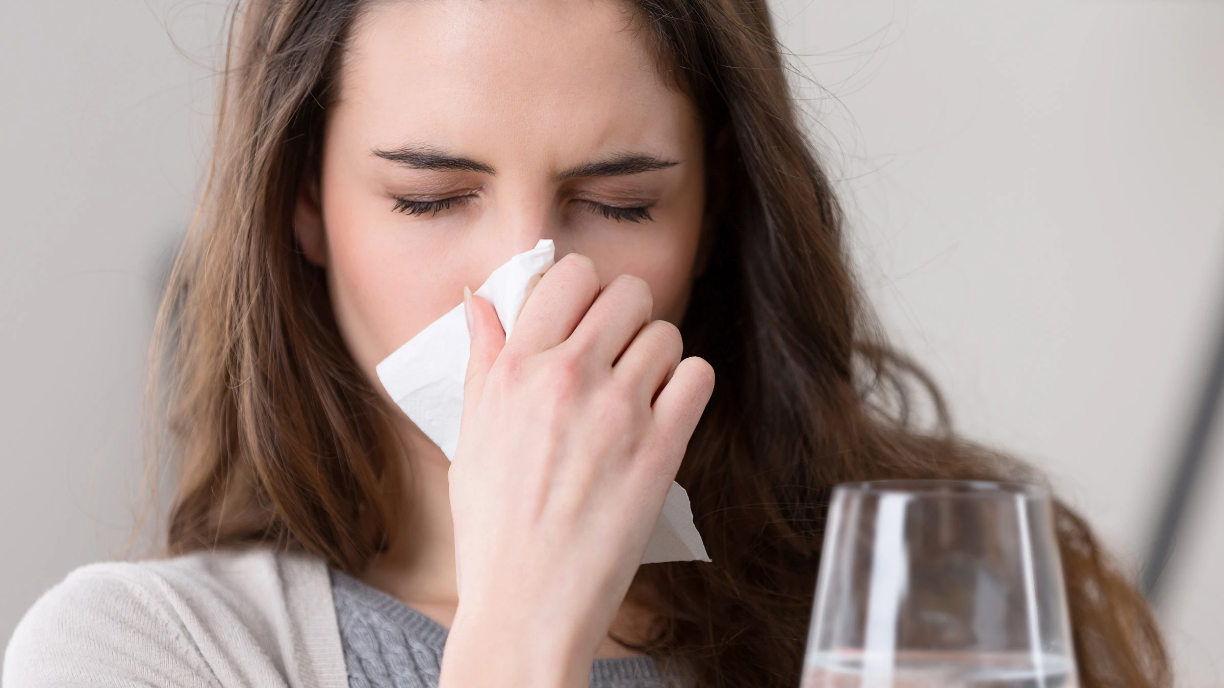 Young woman blowing her nose with a tissue, looking unwell, with a glass of water nearby (staying hydrated)