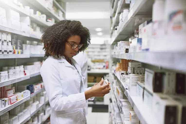 Pharmacist looking for Rx medication price without insurance. The pharmacist is a Black woman wearing a white coat, holding a bottle in her hands.  Shelves of medications surround her in the pharmacy.