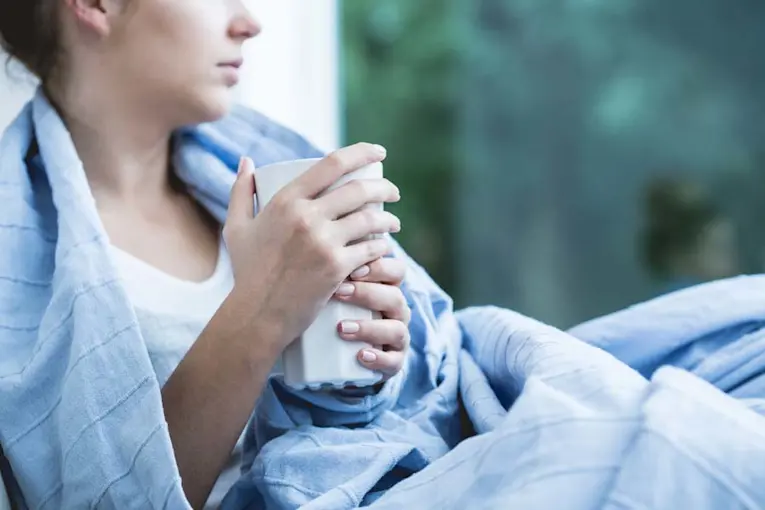 Woman bundled up in a blanket and is holding a warm cup due to having the flu