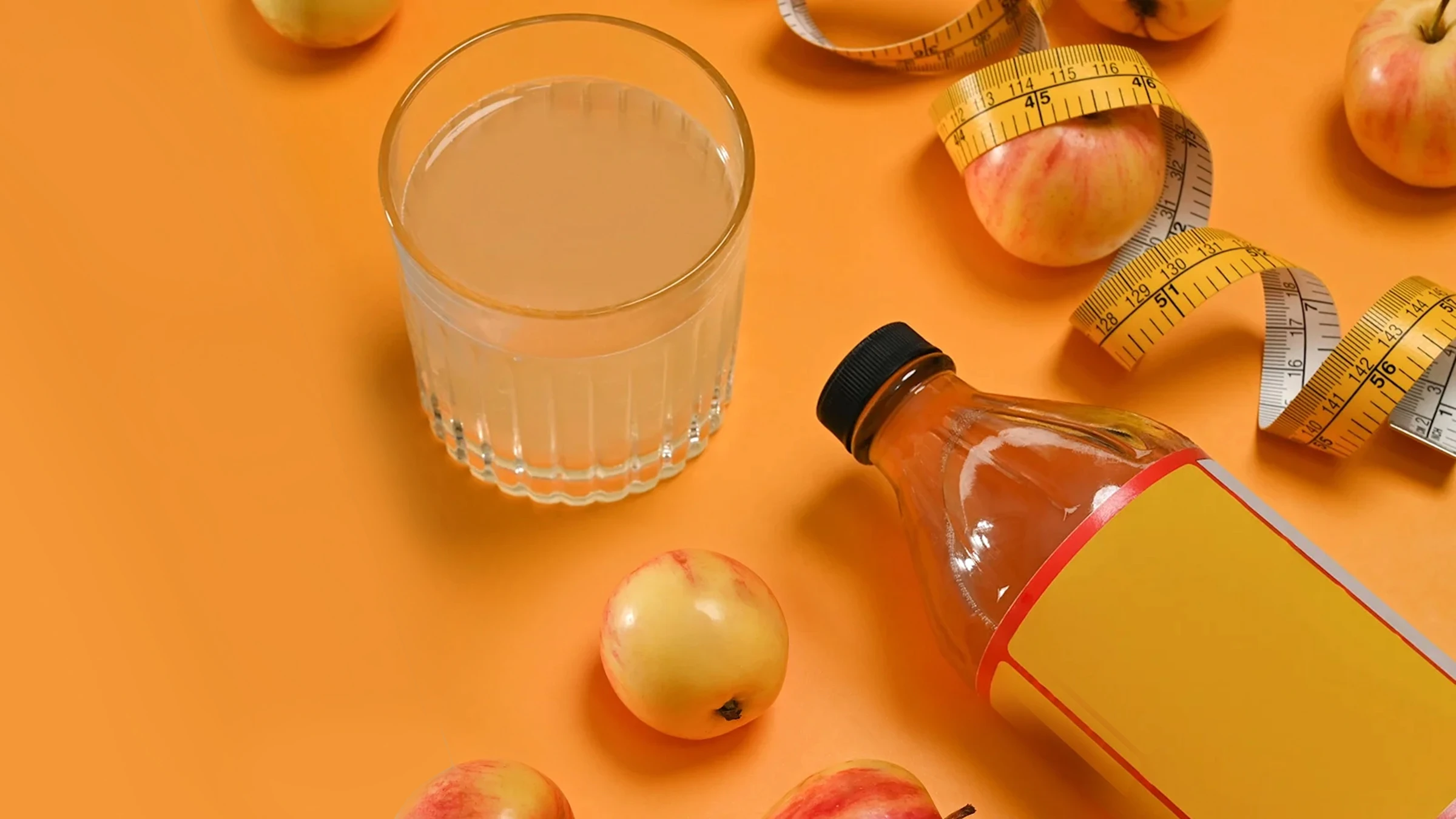 Glass and amber bottle of apple cider vinegar with apples and a yellow measuring tape on orange background