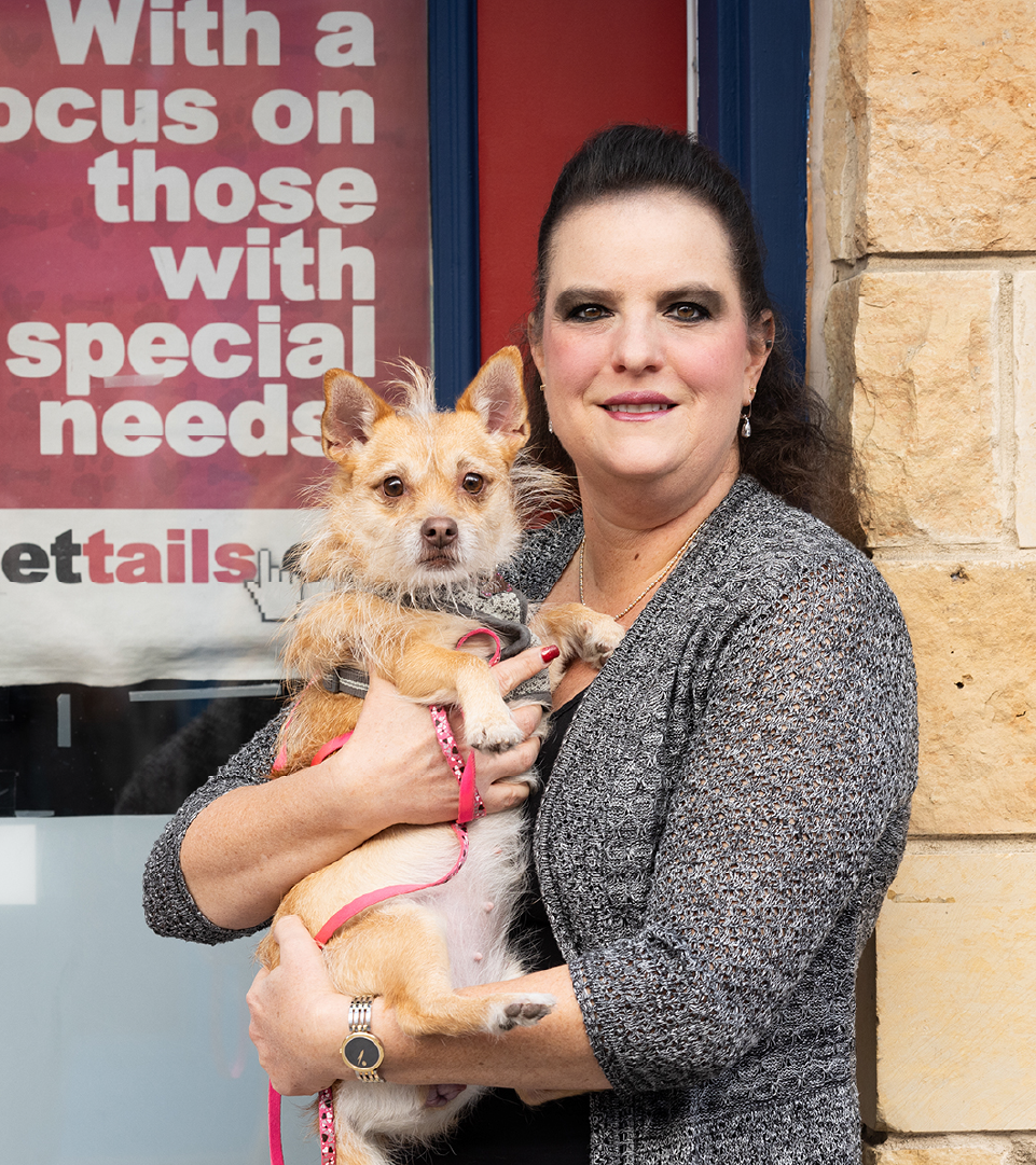 Nicole delaRosa is pictured in front of a red-and-blue door, holding a small tan dog in her arms. A sign on the door reads, “With a focus on those with special needs. streettails.com”